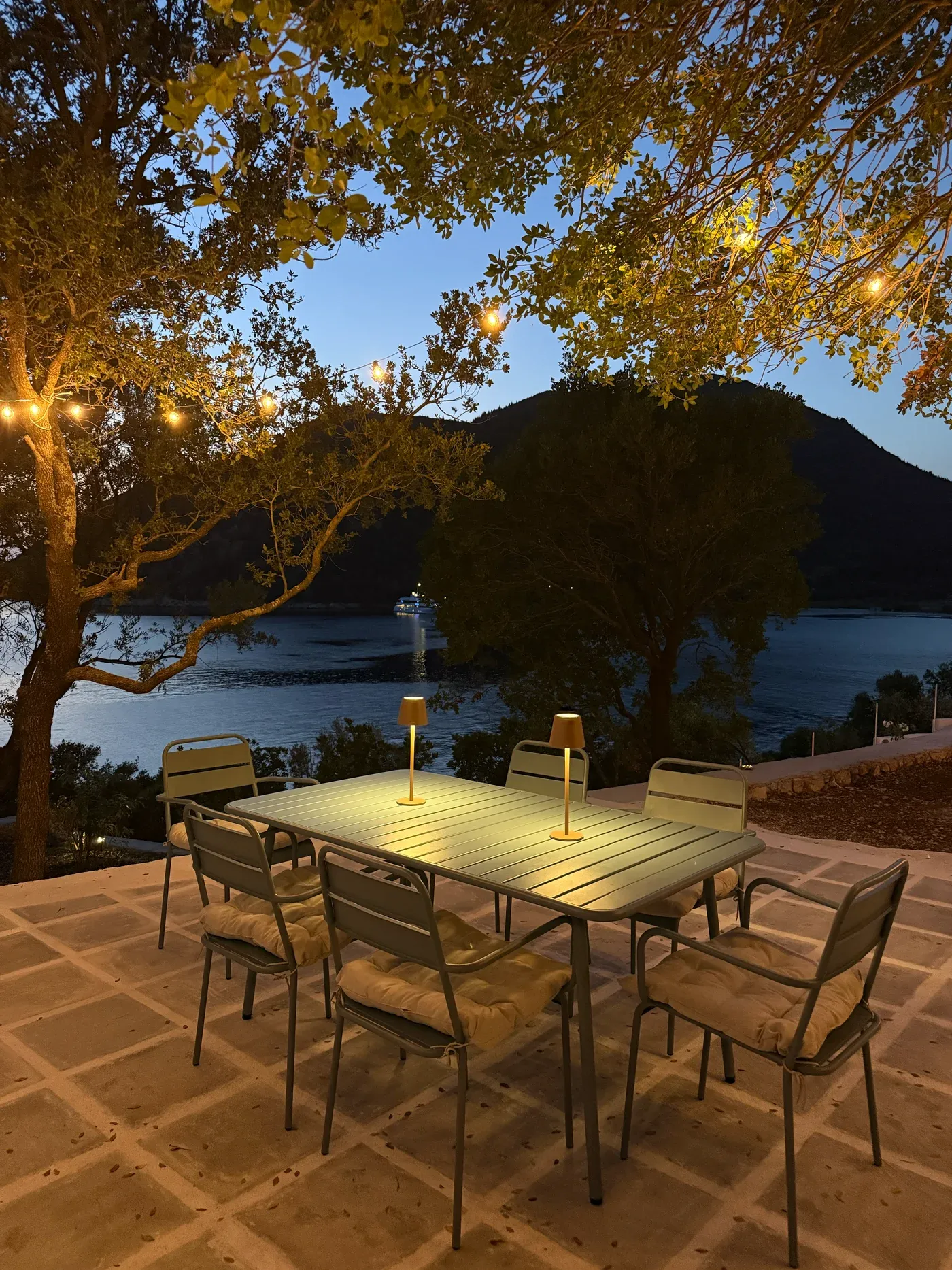 The terrace at dusk: a long table set for six under festoon lights, with the channel and a passing boat below