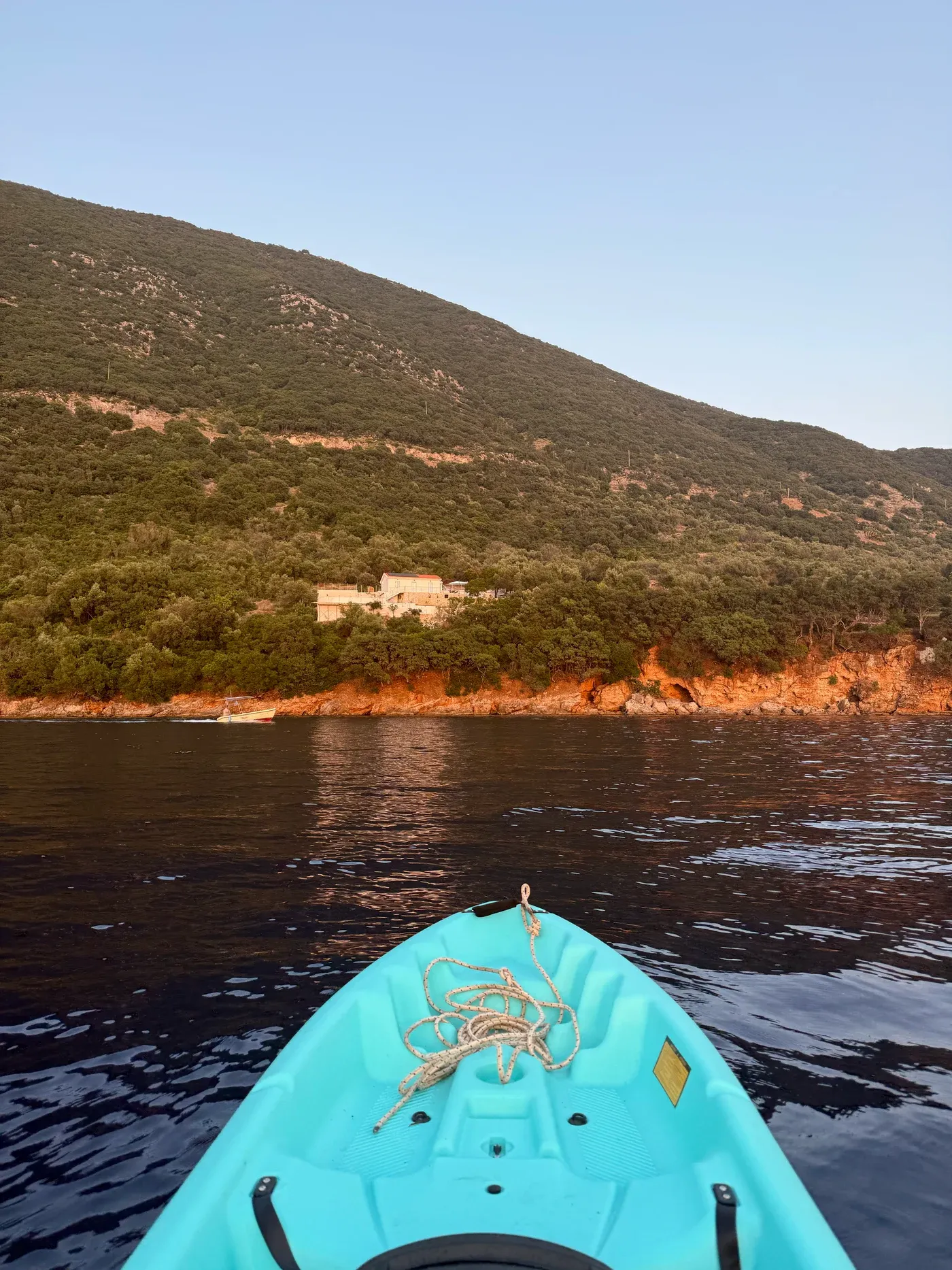 The view from the kayak on flat evening water, looking back at the house among the olive trees above the rocky shore