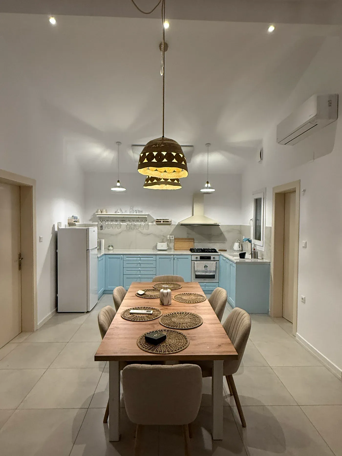 The open kitchen with pale blue cabinets and a long wooden table laid for six under woven pendant lights